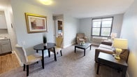 This single-floor unit offers a thoughtfully organized layout that balances comfort and functionality across its key living spaces. The living room is finished with soft wall-to-wall carpeting and painted in a calming light grey tone, creating a relaxed, neutral atmosphere. A window with an exterior view allows natural light to filter in, and a wall-mounted unit provides in-room climate control. A separate dining nook accommodates a small bistro-style table and chairs, adding a defined eating area within the open layout.

The kitchen is compact yet efficiently designed, featuring grey shaker-style cabinetry, laminate countertops with a subtle marble-like pattern, a stainless steel single-basin sink, and a freestanding electric range. Storage is distributed across both upper and lower cabinets.

The bedroom is a standout space, defined by a full exposed brick accent wall that introduces a bold, industrial character to an otherwise soft interior. Carpeted flooring continues throughout, and a window provides an outward view and additional natural light.

The bathroom is clean and straightforward, equipped with a full-size soaking tub and shower combination enclosed by a curtain rod, set against bright white walls and wood-look flooring.