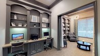 This business center is thoughtfully designed to balance functionality with refined aesthetics. The main workspace area is anchored by a built-in workstation wall finished in a sophisticated charcoal gray, featuring arched cabinetry with decorative pilasters, under-shelf accent lighting, and generous open shelving for display and storage. Multiple desktop computers and a printer are neatly integrated into the continuous countertop, providing ample room for focused work. A glass-paned door with a sidelite panel connects the space to the broader facility, allowing natural light to filter in and maintaining a sense of openness. Adjacent to the workstation area, a private lounge room offers a welcoming retreat, furnished with a pair of upholstered wingback chairs in steel blue leather and a round dark wood coffee table. A large window dressed with patterned teal and gold drapes brings in natural light and an outdoor view, while a striking starburst chandelier overhead adds a touch of elegance. Built-in shelving continues into this room, maintaining design continuity throughout. A curated gallery wall of geometric blue and green artwork adds a finishing touch, creating an environment that is both professional and inviting.