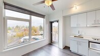 Located at 2180 NE 47th Ave in Portland's vibrant Rose City Park neighborhood, this efficiently designed studio offers a thoughtful single-floor layout that maximizes every square foot. The open-plan living, dining, and kitchen area is bright and airy, featuring large windows dressed with roller shades that flood the space with natural light and frame elevated views of the surrounding Portland neighborhood. Durable wood-look flooring runs throughout the main area, complemented by crisp white walls and a ceiling fan that adds both function and a warm accent. The compact kitchen is well-appointed with sleek light gray cabinetry, a countertop cooktop, an integrated microwave, a sink, and ample upper and lower cabinet storage — all finished in a clean, modern aesthetic. A private balcony accessed directly from the main living area offers open-air views stretching across the Portland skyline and neighborhood rooftops. The entrance provides a defined transition into the living space, while the full bathroom features a walk-in shower with grab bar, a vanity, and a clean white finish. The overall ambiance is contemporary and minimalist, making excellent use of a compact footprint with modern conveniences and abundant natural light.