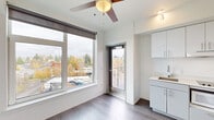 Located at 2180 NE 47th Ave in Portland's vibrant Rose City Park neighborhood, this efficiently designed studio offers a thoughtful single-floor layout that maximizes every square foot. The open-plan living, dining, and kitchen area is bright and airy, featuring large windows dressed with roller shades that flood the space with natural light and frame elevated views of the surrounding Portland neighborhood. Durable wood-look flooring runs throughout the main area, complemented by crisp white walls and a ceiling fan that adds both function and a warm accent. The compact kitchen is well-appointed with sleek light gray cabinetry, a countertop cooktop, an integrated microwave, a sink, and ample upper and lower cabinet storage — all finished in a clean, modern aesthetic. A private balcony accessed directly from the main living area offers open-air views stretching across the Portland skyline and neighborhood rooftops. The entrance provides a defined transition into the living space, while the full bathroom features a walk-in shower with grab bar, a vanity, and a clean white finish. The overall ambiance is contemporary and minimalist, making excellent use of a compact footprint with modern conveniences and abundant natural light.
