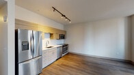 This single-floor unit showcases a contemporary design with a cohesive aesthetic throughout. The galley-style kitchen is well-appointed with a full suite of stainless steel appliances, including a French door refrigerator, electric range, built-in microwave, and dishwasher. Light wood-grain upper and lower cabinetry lines the kitchen wall, complemented by a white subway tile backsplash and light countertops, creating a clean, modern look. Track lighting overhead provides focused illumination across the cooking area.

The open living area flows seamlessly from the kitchen, featuring hardwood-style flooring in a warm, medium-toned finish that runs throughout the unit. The living space benefits from two large windows that frame views of the surrounding urban streetscape, allowing natural light to fill the room. Neutral white walls enhance the brightness and sense of openness.

The bathroom maintains the unit's modern character, featuring a wood-grain vanity with under-cabinet storage, a white countertop with an undermount sink, a framed mirror, and light tile flooring. The overall layout, as seen in the overhead floor plan view, is efficiently organized, connecting the kitchen, living area, and bathroom in a practical and functional arrangement.