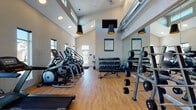 Located at 4528 SW 39th St in Redmond, Oregon, this fitness center is a thoughtfully designed, single-floor facility built to support a full range of exercise routines. The space boasts soaring ceilings that lend an open, airy atmosphere, complemented by an abundance of natural light streaming through tall, strategically placed windows that offer views of the surrounding neighborhood.

The interior features warm hardwood-style flooring throughout, providing both durability and a welcoming aesthetic. A row of cardio equipment — including treadmills, elliptical trainers, and a recumbent bike — lines one side of the room, each machine positioned to take advantage of the natural light. On the opposite side, a well-stocked free weights area offers a comprehensive selection of dumbbells neatly organized on multi-tier racks, accompanied by an adjustable workout bench for strength training.

Floor-to-ceiling mirrors along one wall visually expand the space and allow users to monitor their form during exercise. A vibrant geometric accent wall adds a pop of color and visual interest, while bold pendant lights with black and gold finishes hang from above, enhancing the modern design. A wall-mounted flat-screen TV and posted fitness center guidelines round out the functional amenities. The overall ambiance is clean, energetic, and purpose-built for an effective workout experience.