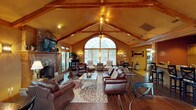 This single-floor space makes a striking impression through its architectural design, centered around a dramatic vaulted ceiling with exposed wooden beam trusses that rise to a generous peak. Warm wood tones throughout the ceiling and structural elements contribute to a cozy yet grand ambiance. A large arched window serves as the focal point of the main living area, framing views of the surrounding greenery while allowing abundant natural light to pour into the space. Flanking the arched window are additional multi-pane windows and glass doors, further enhancing the bright, open feel.

The living area is anchored by a full brick fireplace on one wall, complete with a raised hearth and mantle, providing both a functional and decorative element. Rich hardwood-style flooring runs throughout, complementing the leather seating arranged around a patterned area rug and a glass-top coffee table. Recessed lighting and wall sconces supplement the natural light, maintaining warmth during evening hours. A ceiling fan is centrally mounted at the peak of the vault. The open layout connects the main seating area to additional dining and sitting spaces toward the rear, creating a seamless flow.