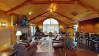 This single-floor space makes a striking impression through its architectural design, centered around a dramatic vaulted ceiling with exposed wooden beam trusses that rise to a generous peak. Warm wood tones throughout the ceiling and structural elements contribute to a cozy yet grand ambiance. A large arched window serves as the focal point of the main living area, framing views of the surrounding greenery while allowing abundant natural light to pour into the space. Flanking the arched window are additional multi-pane windows and glass doors, further enhancing the bright, open feel.

The living area is anchored by a full brick fireplace on one wall, complete with a raised hearth and mantle, providing both a functional and decorative element. Rich hardwood-style flooring runs throughout, complementing the leather seating arranged around a patterned area rug and a glass-top coffee table. Recessed lighting and wall sconces supplement the natural light, maintaining warmth during evening hours. A ceiling fan is centrally mounted at the peak of the vault. The open layout connects the main seating area to additional dining and sitting spaces toward the rear, creating a seamless flow.