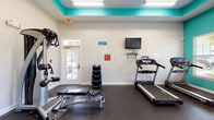 Located at 2350 Freedom Blvd in Florence, South Carolina, this fitness center is a well-equipped, single-floor facility designed to support a full range of workout routines. The space is immediately distinguished by its bold teal accent walls and matching tray ceiling detail, which lend a vibrant, energizing atmosphere to the room. White walls contrast the colorful design elements, keeping the space feeling bright and open.

The flooring throughout is durable rubber matting, ideal for both cardio and strength training activities. Multiple windows fitted with blinds allow generous natural light to fill the room while offering views of the surrounding outdoor greenery. A wall-mounted flat-screen television provides entertainment during workouts.

The cardio section is well-stocked, featuring multiple treadmills, elliptical cross-trainers, and a stair climber, each equipped with individual display consoles. For strength training, the facility includes a multi-function cable machine, an adjustable workout bench, a rack of dumbbells in varying weights, and stability balls. A dedicated station with a sanitizing dispenser and storage cart promotes cleanliness and organization. The space also connects to an adjoining room with hardwood flooring, offering additional utility. A keyed exterior door provides direct outdoor access, adding convenience to this comprehensively outfitted fitness facility.