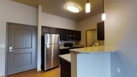 This single-floor space showcases a well-appointed kitchen as its centerpiece, designed with both aesthetics and functionality in mind. The kitchen features rich, dark espresso cabinetry that extends to the upper walls, providing ample storage. Stainless steel appliances, including a full-size refrigerator, built-in microwave, and a black electric range, complement the contemporary design. The light-toned granite countertops provide a striking contrast to the dark cabinetry, adding visual interest and a polished finish.

A peninsula with matching granite extends into the living area, creating a natural division between spaces while offering additional seating and prep space. Pendant lighting hangs above the peninsula, supplemented by a flush-mount ceiling light that brightens the kitchen. The ceilings are notably elevated, contributing to an open, airy atmosphere throughout.

Warm hardwood flooring runs through the space, adding warmth and continuity. A doorway visible from the kitchen leads to what appears to be a bathroom or hallway, indicating a thoughtfully connected floor plan. The entry door is also visible, featuring a classic paneled design.