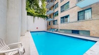 Located at 1745 Avenue Cedar in Montréal, QC, this outdoor pool area offers a well-appointed recreational retreat within an urban setting. The centrepiece is a generously sized swimming pool with vivid blue water, featuring graduated entry steps that provide easy access for swimmers of all levels. The pool is framed by a smooth stone tile deck, offering ample space for lounging and relaxation.

Blue sun loungers are positioned along the poolside, inviting residents to enjoy warm summer days in comfort. The area is enclosed by a combination of clean, light-coloured stucco walls and the warm brick façade of the adjacent multi-storey building, which features glass block accents at its base, balconies above, and large windows that overlook the pool below. This architectural surround creates a semi-private, sheltered atmosphere while still allowing natural light to flood the space.

Lush greenery, including climbing vines and mature trees, softens the hard surfaces and adds a refreshing natural element to the environment. A life ring is mounted on the wall, and pool maintenance equipment is conveniently stored nearby. The overall ambiance is one of a well-maintained, tranquil urban oasis, offering a private escape from the surrounding city.
