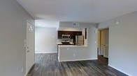 This single-story space features an open-concept design that seamlessly connects the kitchen and living area, creating a cohesive and functional layout. The kitchen is outfitted with dark espresso cabinetry that contrasts sharply against light countertops, providing a modern aesthetic. A peninsula counter extends into the living space, offering additional prep space and serving as a natural divider between the two areas. Stainless steel appliances, including a refrigerator, microwave, and gas range, are integrated throughout the kitchen.

The flooring consists of dark wood-style planks that run continuously across the main living areas, adding warmth and visual continuity. Walls are finished in a neutral light gray tone, complementing the overall modern palette. A textured ceiling is present throughout the space. Recessed lighting and overhead ventilation are visible, ensuring practical functionality. A hallway visible from the main area leads to additional rooms, while a front entryway with a paneled door provides access to the unit. The layout is efficiently designed to maximize the usability of the available space.