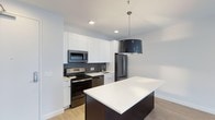 This single-floor 2-bedroom, 2-bathroom residence offers a thoughtfully designed layout with modern finishes and a clean, contemporary aesthetic. The kitchen serves as a focal point of the living space, showcasing crisp white shaker-style cabinetry that extends to the ceiling, maximizing storage. A suite of stainless steel appliances — including a range, microwave, dishwasher, and refrigerator — provides full functionality, while a classic subway-style tile backsplash in a charcoal tone adds a refined contrast to the bright cabinetry. A central kitchen island with a white countertop and dark base offers additional workspace and casual seating potential, anchored by a striking large-drum pendant light overhead. Recessed lighting throughout ensures the space remains bright and well-lit. Light hardwood-style flooring runs underfoot, complementing the neutral white and grey color palette that gives the home a fresh, airy feel. The overall design balances practicality with style, making this residence an appealing option for those seeking modern urban living with well-appointed details and a cohesive, move-in-ready interior.