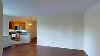 This single-floor space offers a clean, functional layout designed for comfortable everyday living. The main living area is spacious and open, characterized by smooth white walls and rich dark hardwood flooring that extends throughout, creating a warm and cohesive aesthetic. The neutral color palette provides a versatile backdrop, making the space easy to personalize and furnish to a variety of styles.

The living area flows naturally into the kitchen, which is partially separated by a raised breakfast bar counter — an ideal spot for casual dining or entertaining. The kitchen features warm wood cabinetry in a classic finish, adding a touch of traditional character to the space. A stylish cluster pendant light fixture hangs above, casting a warm, inviting glow that enhances the kitchen's ambiance. The countertops and sink are visible from the living area, reinforcing the open and connected feel of the layout.

The overall atmosphere is calm and uncluttered, with the interplay of warm wood tones against crisp white walls and ceilings lending the space a timeless, livable quality. The single-floor plan ensures easy accessibility and a straightforward flow between areas, making this a practical and welcoming space.
