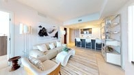 This elegantly designed high-rise residence showcases a contemporary aesthetic throughout, characterized by a neutral palette of whites, creams, and warm wood tones. The open-plan living and dining area is anchored by expansive floor-to-ceiling windows that frame panoramic city views, flooding the space with natural light. The living area features a streamlined entertainment setup, a chrome open shelving unit, and plush seating, while the dining space is distinguished by a solid wood table beneath a striking sculptural gold chandelier and a full-length mirror wall that amplifies the sense of space.

The kitchen is outfitted with dark cabinetry, stainless steel appliances, and a generous waterfall-edge island with bar seating for casual dining. Multiple bedrooms offer soft carpeting, large windows with expansive city views, and private balcony access in select rooms. The primary bedroom includes a generously proportioned upholstered bed and refined furnishings.

Both bathrooms are finished in floor-to-ceiling Calacatta marble, featuring deep soaking tubs, glass-enclosed shower enclosures, floating dark vanities with white countertops, and recessed lighting, creating a spa-like ambiance throughout.