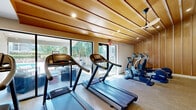 Located in Beaverton, Oregon, this single-floor fitness and wellness facility offers a thoughtfully designed layout spanning a generous footprint. The space is defined by a striking architectural style, with warm wood-paneled walls and elegant sloped wood-plank ceilings that lend a cohesive, natural aesthetic throughout.

The centerpiece of the facility is a spacious, well-equipped exercise room featuring an impressive array of cardio machines — including multiple treadmills, ellipticals, and a recumbent bike — alongside a dedicated strength-training area with a multi-function cable machine, free weights, and resistance equipment. Full-length mirrors maximize the sense of openness, while large floor-to-ceiling windows and glass doors invite abundant natural light and offer views of the outdoor surroundings, including a glimpse of a pool area. An oversized ceiling fan ensures comfortable airflow throughout workouts.

A serene secondary area, finished with light hardwood flooring and full wood-panel walls, provides a calming retreat for stretching or yoga, with a large picture window framing lush greenery. A wall-mounted flat-screen TV adds functionality to the strength training zone.

The bedroom, a generously proportioned private room, features light hardwood floors, cool-toned walls, and a stylish barn-style sliding door. A living room rounds out the accommodations, completing this versatile wellness-oriented space.