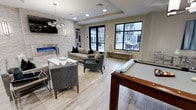 This contemporary single-floor amenity space is thoughtfully designed to serve as a versatile social and recreational hub. The open-plan layout seamlessly connects multiple functional zones, all unified by light wood-style flooring, a neutral color palette, and a clean modern aesthetic.

At the heart of the space sits a full-size pool table, making it an immediate focal point for recreation. Adjacent to it, a comfortable lounge area is anchored by a modern linear electric fireplace set into a striking stacked stone accent wall. A wall-mounted flat-screen TV above the fireplace and a stylish chandelier overhead enhance the entertainment appeal of this seating zone, which features a sofa, accent chairs, and a coffee table arranged for easy conversation.

A separate dining area with a round glass-top table and modern acrylic chairs provides space for casual gatherings. Recessed lighting throughout, complemented by pendant and chandelier fixtures, ensures the space is well-lit at all times.

Large black-framed windows and glass doors flood the interior with natural light and offer clear views of an outdoor patio area, creating a seamless indoor-outdoor connection. The overall ambiance is sleek, bright, and inviting — well-suited for community gatherings, leisure activities, and everyday socializing.