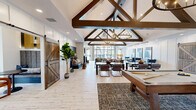 Located at 1062 W Oakland Ave in Johnson City, Tennessee, this single-floor community clubhouse offers a generously sized gathering space designed with a sophisticated yet welcoming aesthetic. The architecture is immediately striking, with dramatic vaulted ceilings supported by bold exposed dark wood trusses and adorned with statement wrought-iron candle-style chandeliers. Whitewashed wood-look tile flooring runs throughout, lending the space a clean, modern farmhouse character.

The main living area is an expansive, light-filled lounge featuring a linear fireplace, a large-screen TV, and an eclectic mix of leather and upholstered seating in warm cognac and rich cobalt tones. Floor-to-ceiling black-framed windows and glass doors flood the room with natural light while offering views of the outdoor pool area. A second living space doubles as a game room, complete with a billiard table and a stylish circular chandelier with decorative metallic orb wall accents.

The kitchen is sleekly outfitted with flat-front wood cabinetry, quartz countertops, a bold navy stacked-tile backsplash, beverage refrigerators, and draft beer taps — ideal for social gatherings. The dining area features built-in booth seating with a contemporary pendant light, offering an intimate setting. A business center lounge with patterned seating and ample natural light rounds out this well-appointed community space.