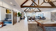 Located at 1062 W Oakland Ave in Johnson City, Tennessee, this single-floor community clubhouse offers a generously sized gathering space designed with a sophisticated yet welcoming aesthetic. The architecture is immediately striking, with dramatic vaulted ceilings supported by bold exposed dark wood trusses and adorned with statement wrought-iron candle-style chandeliers. Whitewashed wood-look tile flooring runs throughout, lending the space a clean, modern farmhouse character.

The main living area is an expansive, light-filled lounge featuring a linear fireplace, a large-screen TV, and an eclectic mix of leather and upholstered seating in warm cognac and rich cobalt tones. Floor-to-ceiling black-framed windows and glass doors flood the room with natural light while offering views of the outdoor pool area. A second living space doubles as a game room, complete with a billiard table and a stylish circular chandelier with decorative metallic orb wall accents.

The kitchen is sleekly outfitted with flat-front wood cabinetry, quartz countertops, a bold navy stacked-tile backsplash, beverage refrigerators, and draft beer taps — ideal for social gatherings. The dining area features built-in booth seating with a contemporary pendant light, offering an intimate setting. A business center lounge with patterned seating and ample natural light rounds out this well-appointed community space.