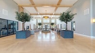 Located at 3223 W 111th St in Naperville, IL, this impressive single-floor clubhouse spans over 5,300 square feet of thoughtfully designed community space. The grand entrance lobby commands immediate attention with its soaring two-story ceilings, exposed wood beam coffered detailing, and a striking central tiered fountain. Mature indoor trees in contemporary planters flank the open space, while a statement brass chandelier adds warmth and elegance. Polished concrete floors and abundant natural light from clerestory windows complete the refined atmosphere.

The living room features vaulted ceilings, floor-to-ceiling black-framed windows that frame outdoor pool views, and a floor-to-ceiling whitewashed stone fireplace with a mounted television. A second lounge area offers comfortable seating with direct sightlines to the pool and landscaped surroundings.

The kitchen and dining area are outfitted with light wood cabinetry, dark stone countertops, brass fixtures, herringbone tile backsplash, open shelving, and stainless steel appliances, with adjacent dining seating bathed in natural light.

The fully equipped fitness center includes treadmills, ellipticals, multi-function strength machines, vaulted beam ceilings, ceiling fans, wall-mounted televisions, and large windows overlooking the outdoors. Multiple connecting hallways tie each area together seamlessly.