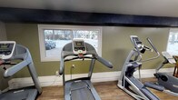 This single-floor community amenity space is thoughtfully designed to serve both fitness and laundry needs in one convenient location. The fitness area is outfitted with cardio equipment including treadmills, elliptical trainers, and a stationary bike, providing a well-rounded workout experience. Olive green accent walls, wood-look flooring, and a bold dark overhead beam give the gym area a distinctive, grounded aesthetic. Multiple windows line the walls, allowing natural light to filter in and offering views of the surrounding outdoor area. Flush-mounted overhead lighting ensures the space remains bright and functional. A casual seating nook with a wooden table and chairs offers a comfortable spot to rest, and a vending machine is conveniently located nearby. An adjacent dedicated weight and strength training room provides additional workout options. The expansive laundry room features rows of washers and dryers arranged along both sides of a central folding island with a clean white countertop surface, making laundry tasks efficient and organized. The neutral gray walls, wood-look flooring, and recessed lighting create a clean, utilitarian atmosphere throughout. Together, these amenities form a practical and well-maintained facility designed to support everyday community living.