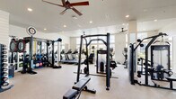 Located at 1841 Dusty Miller Lane in Wilmington, NC, this single-floor fitness center is a comprehensively outfitted facility designed to support a wide range of workout routines. The space features a bright, open layout with high ceilings accented by recessed lighting and ceiling fans that maintain a comfortable environment throughout. Large multi-pane windows line the walls, flooding the interior with natural light and offering views of the surrounding greenery, contributing to an airy and motivating atmosphere. The interior walls are finished in crisp white with board-and-batten detailing, giving the space a clean, modern aesthetic. The facility is divided into dedicated zones for both strength training and cardiovascular exercise. The strength area is stocked with a robust selection of selectorized cable machines, a functional trainer rig loaded with accessories including battle ropes and medicine balls, a multi-station cable system, plate-loaded machines, and a dumbbell rack. The cardio section lines one side of the space with a row of treadmills and upright exercise bikes. Full-length mirrors are strategically placed throughout to enhance spatial perception and support proper form during workouts. Additional amenities include a sanitation station and a small refreshment area, making this fitness center a thorough and user-focused training environment.