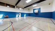 Located at 301 T Street in Tumwater, Washington, this indoor basketball court is a well-appointed, single-floor recreational facility designed to accommodate a variety of court sports. The space is defined by its soaring ceiling height, which is accentuated by bold, dark exposed beams running across the upper expanse, creating an open and airy atmosphere throughout.

Natural light floods the court through a series of skylights positioned along the roofline, supplemented by overhead fluorescent fixtures to ensure consistent illumination at all hours. The walls feature a two-tone paint scheme — a crisp white upper half and a bold royal blue lower half — which adds a vibrant, athletic character to the space while also providing functional wall padding for player safety.

The polished concrete floor is lined with multiple overlapping court markings in black, red, and yellow, supporting a range of sports including basketball, volleyball, and other multi-sport uses. Two wall-mounted basketball backboards with regulation hoops are positioned at each end of the court. Volleyball net pole bases are also visible, highlighting the court's multi-sport versatility.

Upper-level observation windows provide a view into the court from an adjacent space, and multiple access doors are located along the perimeter walls, ensuring convenient entry and exit for players and spectators alike.