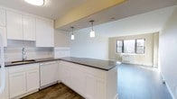 This single-floor space showcases a thoughtfully designed open-concept layout that seamlessly connects the kitchen and living area. The kitchen is outfitted with crisp white shaker-style cabinetry, a dark countertop peninsula that doubles as a breakfast bar, and white appliances including a dishwasher. A subway tile backsplash adds a clean, classic detail to the cooking space. Two pendant lights hang above the peninsula, providing focused task lighting while enhancing the visual transition between the kitchen and the adjoining living area. The flooring throughout is a rich, dark hardwood, lending warmth and continuity to the space. The living area benefits from natural light streaming through a pair of windows, offering views of surrounding greenery. A baseboard heating unit is positioned beneath the windows. The walls are painted in a neutral palette of whites and soft warm tones, creating a bright and welcoming ambiance. The open flow between spaces makes the overall layout feel spacious and functional, well-suited for both everyday living and entertaining.