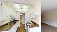 This single-floor residence offers a well-organized and functional layout throughout. The galley-style kitchen is a standout feature, showcasing white cabinetry that lines both sides of the corridor, complemented by dark granite-style countertops and durable wood-look flooring. A double basin sink, gas range with oven, refrigerator, and overhead microwave provide all essential appliances, while a distinctive skylight-style panel ceiling adds a bright, airy feel to the cooking space. A pass-through connects the kitchen to an additional utility area beyond.

The living area is generously sized, finished with neutral-toned carpet and warm beige walls that create a welcoming atmosphere. Vertical blinds cover a large window, allowing for ample natural light and outdoor views, while a sliding glass door provides convenient access to the exterior.

The bedroom continues the same soft carpeting and neutral palette, with a window fitted with vertical blinds that bring in natural light. The space is clean and versatile, offering a comfortable and quiet retreat.

The bathroom features a lengthy vanity with white cabinetry, a single sink set into a dark countertop, and dual mirrors. A separate water closet houses the toilet and a full-size tub and shower combination with clean white tile surround, completing this practical and well-appointed residence.