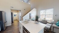 This two-story home showcases a thoughtfully designed interior that balances modern finishes with an open, airy feel. The main level centers around a well-appointed kitchen featuring white shaker cabinetry, quartz countertops, stainless steel appliances including a range, microwave, and refrigerator, and a central island with an integrated sink. Dark hardwood flooring runs throughout the main level, grounding the space with warmth and contrast. A living area adjacent to the kitchen benefits from large windows dressed with white blinds, flooding the space with natural light and offering views of the surrounding neighborhood.

A staircase leads to the upper level, where dramatic vaulted ceilings with recessed lighting create a striking sense of volume. The primary bedroom is generously proportioned, featuring soaring angled ceilings, a walk-in closet with built-in shelving, and a seating area. Additional bedrooms on both floors are carpeted and well-lit by multiple windows. Two full bathrooms serve the home, each finished with white vanities, framed mirrors, and wood-look tile flooring. The overall aesthetic is clean and contemporary, with a neutral color palette that creates a cohesive, light-filled environment throughout.