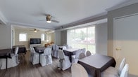 This single-floor space is thoughtfully arranged to accommodate events and gatherings, combining a formal dining area with a connected lounge section. The main dining area features rectangular tables dressed in dark linens, surrounded by chairs adorned with white satin covers and gray sashes, lending an elegant and polished atmosphere. Large vertical-blind windows line one wall, allowing generous natural light to fill the room while offering views of the surrounding greenery outdoors. Crown molding frames the ceiling, adding a refined architectural detail throughout the space. Ceiling fans are installed in both areas, contributing to comfort during events. The adjacent lounge area features a large flat-screen television, a floor lamp, and additional seating, providing a relaxed setting that complements the more formal dining section. Wood-look flooring runs throughout, tying both spaces together cohesively. The neutral gray and white color palette creates a versatile backdrop suitable for a variety of event styles, from intimate gatherings to more structured celebrations. Overall, the space offers a functional and welcoming environment well-suited for hosting events.