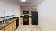 This single-floor residential unit offers a straightforward, functional layout designed for comfortable everyday living. The open-concept living and dining area is finished with neutral-toned wall-to-wall carpeting and features a dedicated entryway with a full-length door. Natural light filters into the space through windows fitted with blinds, lending a bright and airy feel throughout.

The kitchen is positioned adjacent to the living area, separated by a raised breakfast bar that provides both a casual dining option and a natural division between spaces. It is equipped with oak-toned cabinetry, dark granite-style countertops, and black appliances including a range, microwave, refrigerator, and dishwasher. Light tile flooring adds a clean, practical finish to the kitchen area.

The bedroom is generously sized with soft carpeting underfoot, a ceiling fan with integrated lighting, and a window with blinds for natural light and privacy. A closet provides built-in storage within the room.

The bathroom includes a full bathtub and shower combination with a curtain rod, a single vanity with a granite-style countertop, an undermount sink, and a well-lit mirror above. The unit also includes a dedicated in-unit laundry area, pre-fitted with washer and dryer hookups, a dryer vent, and an overhead wire shelving unit for added storage convenience.