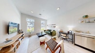 This thoughtfully designed one-bedroom, one-bathroom residence at 1 Gibson Way in Revere, MA offers a bright, contemporary living experience on a single floor. The open-concept kitchen and living area serves as the heart of the home, featuring light-toned hardwood flooring, recessed lighting, and crisp white walls that amplify the natural light streaming in through large windows. The kitchen is outfitted with warm wood-grain cabinetry, stainless steel appliances including a refrigerator, range, microwave, and dishwasher, complemented by a white subway tile backsplash and open shelving for added character. A dining area with a rounded pedestal table connects seamlessly to the kitchen and living space. The living room offers generous window views of the surrounding community, creating an inviting, airy atmosphere. The bedroom is a calm, comfortable retreat with a large window, soft carpeting, and a dedicated closet. The bathroom features a glass-enclosed walk-in shower with neutral tile surround and polished chrome fixtures. Additional conveniences include an in-unit laundry and a utility room, rounding out this well-appointed and efficiently organized home.