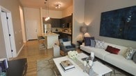 Located at 704 15th St in Durham, NC, this single-floor residence showcases a thoughtfully designed open-concept layout that seamlessly connects the kitchen, dining, and living areas. The kitchen is outfitted with dark cabinetry, stainless steel appliances including a refrigerator, microwave, and dishwasher, and a light-toned granite-topped island with bar seating — an ideal setup for both meal preparation and casual dining. Pendant lighting with warm-toned bulbs hangs above the island, adding an inviting ambiance to the space. The living area is generously proportioned, featuring neutral-toned walls that complement the warm hardwood-style flooring found throughout. The space is styled with a contemporary aesthetic, highlighted by a plush sofa, accent chairs, a marble-patterned area rug, and a sleek white coffee table. Soft lighting from floor and table lamps enhances the warm, welcoming atmosphere. The open floor plan creates a strong sense of connectivity between all areas, making the space feel expansive and well-suited for both everyday living and entertaining.