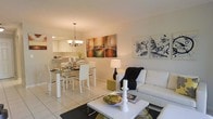 This single-floor space showcases an open-concept design that integrates the living, dining, and kitchen areas into one cohesive, flowing environment. Light-toned ceramic tile flooring runs throughout, lending a clean and bright aesthetic to the interior.

The living area is appointed with a white sofa accented by a dark throw blanket, paired with a contemporary white coffee table. Abstract wall art adds visual interest and personality to the neutral walls. A black table lamp provides ambient lighting, complementing the overall modern aesthetic.

Transitioning naturally into the dining area, a glass-top dining table surrounded by white chairs serves as a stylish and functional centerpiece. An elegant chandelier hangs overhead, providing warm, focused lighting that enhances the dining experience.

The kitchen, visible through a pass-through opening, features white cabinetry and ample counter space, maintaining the clean and cohesive color palette established throughout the space. The open sightlines between all three areas create a sense of spaciousness, making the layout ideal for both everyday living and entertaining. The overall ambiance is modern, airy, and well-coordinated.