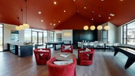 Located at 17156 SW Woodpecker Ln in Beaverton, Oregon, this single-floor community clubroom delivers a bold and dynamic social environment designed for gathering and recreation. The space is immediately defined by its dramatic vaulted ceiling, painted a deep, rich red that draws the eye upward and sets a vibrant tone throughout. Large floor-to-ceiling windows line the perimeter, flooding the interior with natural light and offering expansive views of the surrounding Beaverton landscape.

The open layout is thoughtfully divided into distinct activity zones. A lounge area anchors the space with a generous sectional sofa, accent chairs, and a low coffee table arranged atop a dark area rug — ideal for relaxed conversation. Nearby, a cluster of bold red upholstered barrel chairs surrounds a round game table, providing a cozy yet lively setting. Recreation is well-served with both a pool table and a shuffleboard table positioned throughout the room.

A built-in bar and dining area features bar-height seating, a large flat-screen display mounted on a striking dark chevron-patterned accent wall, and globe pendant lighting overhead. The adjacent kitchenette includes dark cabinetry, a metallic tile backsplash, under-counter appliances, and a freestanding island — fully equipped for hosting events. Globe pendant lights suspended at varying heights complete the polished, modern aesthetic.