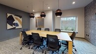 Located at 701 5th Avenue North in Seattle, WA, this professional meeting and collaborative space is thoughtfully designed to accommodate a variety of work styles and group sizes. The centerpiece is a spacious boardroom enclosed by a striking black-framed glass partition wall, offering both visual transparency and acoustic separation. The boardroom features a large rectangular wood conference table surrounded by ergonomic black task chairs, illuminated overhead by a series of dramatic copper and black globe pendant lights. A window along one wall invites natural light and provides a view of the exterior greenery. Contemporary artwork adorns the walls, adding a creative touch to the professional setting. Adjacent to the boardroom is a comfortable lounge area appointed with warm cognac leather armchairs, a round coffee table, and a textured, patterned feature wall with circular sconce lighting. The lounge offers an inviting space for informal conversations or breakout discussions. Additional seating areas with task chairs and small tables are arranged throughout the open layout, providing flexible options for individual work or small group collaboration. Patterned carpet flooring with teal accent lines ties the space together, while recessed lighting and pendant fixtures maintain a warm, polished ambiance throughout.