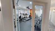 This fitness center offers a comprehensive workout environment spread across a single floor, thoughtfully designed to accommodate a wide range of exercise needs. The main workout area is bright and inviting, featuring large windows that allow natural light to fill the space while providing views of the surrounding exterior. A bold orange accent wall adds a dynamic, energizing touch to the otherwise clean, white interior.

The cardio zone is well-stocked with multiple treadmills, elliptical trainers, and stationary and recumbent bikes, all positioned to take advantage of the natural light. Overhead, ceiling-mounted fans ensure comfortable airflow throughout workout sessions, complemented by recessed lighting for consistent illumination. A wall-mounted television is also present, keeping users entertained during cardio sessions.

A dedicated strength training area houses a multi-function cable machine, a weight bench, and a full rack of dumbbells displayed on tiered shelving, catering to users of all fitness levels. A separate room provides additional space for targeted strength work, including a punching bag and a step platform for varied training routines.

Durable rubber flooring throughout the facility provides safety and sound absorption, while the clean, organized layout ensures ease of movement between equipment. Glass-paneled entry doors lend a polished, professional aesthetic to the overall space.
