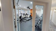 This fitness center offers a comprehensive workout environment spread across a single floor, thoughtfully designed to accommodate a wide range of exercise needs. The main workout area is bright and inviting, featuring large windows that allow natural light to fill the space while providing views of the surrounding exterior. A bold orange accent wall adds a dynamic, energizing touch to the otherwise clean, white interior.

The cardio zone is well-stocked with multiple treadmills, elliptical trainers, and stationary and recumbent bikes, all positioned to take advantage of the natural light. Overhead, ceiling-mounted fans ensure comfortable airflow throughout workout sessions, complemented by recessed lighting for consistent illumination. A wall-mounted television is also present, keeping users entertained during cardio sessions.

A dedicated strength training area houses a multi-function cable machine, a weight bench, and a full rack of dumbbells displayed on tiered shelving, catering to users of all fitness levels. A separate room provides additional space for targeted strength work, including a punching bag and a step platform for varied training routines.

Durable rubber flooring throughout the facility provides safety and sound absorption, while the clean, organized layout ensures ease of movement between equipment. Glass-paneled entry doors lend a polished, professional aesthetic to the overall space.