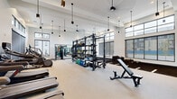 Located at 4607 Xenia St in Denver's Central Park neighborhood, this well-appointed fitness center offers a spacious, single-level layout designed to accommodate a full spectrum of workout needs. The facility's most striking architectural feature is its soaring ceiling height, accentuated by floor-to-ceiling windows and clerestory glazing that flood the interior with natural light and provide open views of the surrounding neighborhood. Sleek pendant lighting fixtures are suspended from above, complementing the clean, modern aesthetic defined by a crisp white palette and dark-trimmed window frames.

The main workout floor is divided into distinct functional zones. A dedicated strength and functional training area features a large multi-station rig stocked with barbells, weight plates, medicine balls, battle ropes, resistance bands, and a punching bag. An expansive rubber-floored turf zone with marked lanes provides ample space for agility drills, sled work, and dynamic exercises. The cardio section is equipped with stair climbers, ellipticals, treadmills, and rowing machines. Full-length mirrors are strategically positioned throughout to maximize spatial awareness. A separate corridor leads to additional support spaces, completing this comprehensively equipped facility.