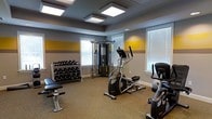 Located at 1518 E Park Ave in Savannah, Georgia, this single-floor fitness center is a well-appointed exercise facility designed to support a comprehensive workout experience. The space features a thoughtfully arranged layout that maximizes functionality while maintaining an open, accessible feel.

The interior is finished with neutral gray carpeting underfoot and walls painted in a bold combination of gray and yellow horizontal stripes, lending the room an energetic and motivating atmosphere. Flush-mounted square light panels in a coffered ceiling section provide bright, even illumination throughout the space.

The facility is equipped with a variety of cardio and strength training equipment, including an elliptical trainer and a stationary cycle for cardiovascular workouts. A multi-cable functional training station serves as a central strength training anchor, offering a wide range of resistance exercises. A full dumbbell rack stocked with multiple weight sets is positioned along the wall alongside a dedicated weight bench, making the space suitable for both beginners and experienced fitness enthusiasts.

Multiple windows fitted with white horizontal blinds allow ample natural light to fill the room while providing privacy control. The overall ambiance is clean, practical, and well-maintained, making this fitness center a convenient and capable facility for residents or visitors seeking a reliable place to exercise.
