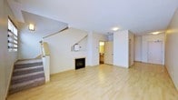 This two-floor residence offers a thoughtfully designed layout that balances openness with functional living spaces. The main level is anchored by a bright, open living area finished with light-toned hardwood flooring that extends throughout, lending the space a clean and airy feel. A fireplace with a simple surround serves as a focal point in the living room, adding warmth and character to the space. A staircase with carpeted treads and a wood railing leads to the upper floor, positioned along one wall to maximize the usable floor area below. Adjacent to the living space, a glimpse into the kitchen reveals cabinetry in a warm wood finish, with access to what appears to be additional utility or storage areas nearby. A secondary door on the main level offers convenient access, likely to an exterior entry or closet. The neutral white walls and flush ceiling lighting throughout keep the interiors feeling fresh and versatile. The upper level provides additional living or sleeping accommodations, completing the functional two-story layout. Overall, the space presents a practical and welcoming environment with classic finishes well-suited for everyday living.
