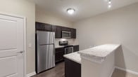Located at 40 Hutchinson Ave in Columbus, Ohio, this single-floor residence offers a thoughtfully designed layout with modern finishes and practical features throughout. The kitchen is a standout space, equipped with stainless steel appliances including a refrigerator, electric range, and over-the-range microwave. Rich dark cabinetry contrasts elegantly with light stone-look countertops, while a classic subway tile backsplash adds a polished touch. A peninsula counter provides additional workspace and a natural transition into the living areas. Hard-surface flooring runs through the kitchen and utility areas, lending a clean, contemporary look. The living area is generously sized and finished with plush carpeting, creating a warm and comfortable environment with natural light from a nearby window. The bathroom features a spacious walk-in shower with sliding glass doors, built-in shelving, and dark wood-toned vanity cabinetry that mirrors the kitchen's aesthetic. Storage is a notable strength of this residence, with multiple walk-in closets outfitted with wire shelving systems offering ample organization options. A dedicated laundry room houses a full-size washer and dryer alongside additional shelving. The overall design maintains a consistent neutral color palette, clean white trim, and a calm ambiance throughout.