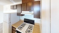 This single-floor unit offers a straightforward and functional layout designed for comfortable everyday living. The living area is generously proportioned, featuring wall-to-wall carpet that adds warmth underfoot, neutral-toned walls, and a textured ceiling. Natural light filters in through windows fitted with blinds, brightening the open space. A second carpeted room provides a quiet, private retreat, also benefiting from natural light through a wide window.

The kitchen is equipped with a gas range and overhead range hood, complemented by dark wood-toned upper cabinetry that provides ample storage. A full-size refrigerator is also present, and the kitchen connects conveniently to the main living area. The transition between rooms is marked by a tiled entryway near the front door, offering easy access and a clear delineation of spaces.

The bathroom is well-appointed with a dual-sink vanity topped with a light countertop, a large wall mirror above, and a combination tub and shower enclosed in tiled surround. A towel bar and ventilation fan complete the practical fixtures. Throughout the unit, the design is clean and utilitarian, providing a neutral canvas well-suited for personalization.