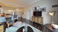 This well-appointed one-bedroom, one-bathroom residence is thoughtfully designed across a single floor, offering a seamless blend of modern aesthetics and functional living. The open-concept layout connects the kitchen, dining, and living areas, creating a cohesive and inviting atmosphere throughout.

The kitchen is outfitted with sleek white cabinetry, stainless steel appliances, a mosaic tile backsplash, dark countertops, and pendant lighting that adds warmth to the space. Dark hardwood flooring runs through the main living areas, providing a rich contrast to the lighter walls and furnishings.

The living room is bathed in natural light through floor-to-ceiling windows and offers direct access to a private balcony with urban views. The space is open and airy, with room for comfortable seating and dining arrangements.

The bedroom features plush carpeting, cool blue-gray walls, and a standout intricately carved wood headboard that anchors the room. A large window with sheer curtains allows soft natural light to filter in.

The bathroom is finished with large-format white tile flooring, wood-toned cabinetry, and a clean, contemporary design that completes this polished urban residence.