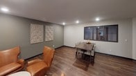 This single-floor space is thoughtfully organized into distinct functional areas, each designed to serve the needs of its occupants. The common lounge area features warm wood-look flooring, neutral-toned walls accented with decorative wall art panels, and comfortable seating including a tan leather accent chair. Recessed lighting illuminates the space evenly, creating a welcoming atmosphere. An interior window connects the lounge to an adjacent room, adding a sense of openness.

The dining area, positioned within the same open zone, accommodates a dark wood dining table surrounded by woven upholstered chairs, making it suitable for group gatherings. Decorative geometric wall panels add a subtle design element to the space.

A separate laundry facility is equipped with multiple commercial-grade Speed Queen front-load washers and dryers, along with a utility sink and a large folding table for convenience. Instructional signage is posted for ease of use. A window in the laundry room provides natural light. The consistent use of wood-look flooring and neutral wall colors throughout ties the various areas together into a cohesive, functional environment.