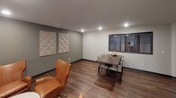 This single-floor space is thoughtfully organized into distinct functional areas, each designed to serve the needs of its occupants. The common lounge area features warm wood-look flooring, neutral-toned walls accented with decorative wall art panels, and comfortable seating including a tan leather accent chair. Recessed lighting illuminates the space evenly, creating a welcoming atmosphere. An interior window connects the lounge to an adjacent room, adding a sense of openness.

The dining area, positioned within the same open zone, accommodates a dark wood dining table surrounded by woven upholstered chairs, making it suitable for group gatherings. Decorative geometric wall panels add a subtle design element to the space.

A separate laundry facility is equipped with multiple commercial-grade Speed Queen front-load washers and dryers, along with a utility sink and a large folding table for convenience. Instructional signage is posted for ease of use. A window in the laundry room provides natural light. The consistent use of wood-look flooring and neutral wall colors throughout ties the various areas together into a cohesive, functional environment.