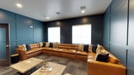 This single-floor space makes a strong design statement through its bold, dark teal paneled accent walls, which lend a sense of depth and refined elegance to the interior. Recessed ceiling lights are evenly distributed throughout, providing warm, focused illumination that complements the room's sophisticated color palette. The centerpiece of the space is a large, curved cognac leather sectional sofa adorned with an assortment of decorative pillows in navy, gold, and textured tones — offering generous seating capacity and a luxurious, inviting feel. The sectional is positioned around a low-profile coffee table, creating a natural gathering area suited for relaxed socializing or lounging. Neutral roller shades dress the windows, which allow diffused natural light to soften the darker wall tones. The flooring features a patterned carpet tile in muted, earthy tones that grounds the space and adds subtle texture underfoot. Ceiling-mounted HVAC vents and in-ceiling speakers suggest the space is thoughtfully equipped for both comfort and entertainment. Overall, the space balances a modern, upscale aesthetic with a comfortable, functional layout ideal for communal use.