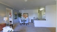This single-story space showcases an open-concept design that seamlessly connects the dining area and kitchen into one cohesive living environment. The layout promotes a sense of spaciousness, with the dining zone anchored by a round glass-top table surrounded by upholstered chairs in a soft gray tone. Decorative touches, including a large framed mirror, abstract wall art, and a tall indoor plant with vibrant orange florals, add warmth and personality to the neutral color palette of gray and white walls.

The kitchen is positioned along one side of the space, featuring white cabinetry, dark countertops, and a peninsula with an integrated sink that serves as both a functional workspace and a subtle divider between the kitchen and dining areas. Standard appliances, including a refrigerator, are neatly integrated into the cabinetry layout. Recessed and overhead lighting illuminate the kitchen efficiently, while a ceiling fan overhead provides comfort throughout the open area. Plush wall-to-wall carpeting runs throughout, contributing to a cozy and comfortable atmosphere overall.