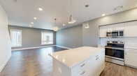 This contemporary 3-floor home is designed with a clean, modern aesthetic carried consistently throughout each level. The main living area features an open-concept layout where the kitchen flows seamlessly into the living space. Wide-plank hardwood-style flooring anchors the area, complemented by soft gray walls and recessed lighting paired with decorative pendant fixtures. A central kitchen island with a quartz countertop serves as a focal point, while the kitchen itself is outfitted with white flat-panel cabinetry, a subway tile backsplash, stainless steel appliances including a range, microwave, refrigerator, and dishwasher, and generous counter space. A staircase connects the floors, maintaining the home's bright, open feel. A powder room on the main level offers a pedestal sink and wood-look flooring. Upper-level bedrooms feature plush carpeting, ceiling fans, and natural light from windows, alongside walk-in closets with built-in hanging rods. The full bathroom includes large-format tile flooring, a single vanity with quartz countertop, a round mirror, and a walk-in shower. Throughout, the home's neutral palette and thoughtful finishes create a cohesive, move-in-ready environment.