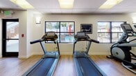 This fitness center offers a comprehensive workout environment designed to accommodate a wide range of exercise routines. The space features warm hardwood flooring throughout, a drop ceiling with recessed and panel lighting, and large windows that provide views of an outdoor pool area, filling the room with natural light and a sense of openness.

The cardio section is equipped with Matrix treadmills and an elliptical trainer, positioned near the windows to allow exercisers to enjoy the outdoor view. A central multi-functional cable training station serves as the focal point of the strength training area, offering versatile resistance options for full-body workouts. A dedicated free weights section includes a full dumbbell rack with a wide weight range, complemented by a SPRI kettlebell rack with color-coded kettlebells in various weights.

Additional functional fitness accessories include medicine balls and stability balls, neatly organized on a storage rack. Floor-to-ceiling mirrors line one wall, enhancing the sense of space and allowing users to monitor their form. A distinctive burnt-orange accent ceiling detail adds a modern, energetic visual element to the space. Glass panel doors connect to an adjoining room, providing additional flexibility. Framed fitness-themed artwork decorates the walls, contributing to the motivating atmosphere of this well-appointed facility.