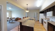 This single-floor residential unit offers a functional and cohesive layout designed for comfortable shared living. The kitchen is a standout feature, showcasing rich dark cabinetry, granite countertops, and light hardwood flooring. A large center island with a built-in sink and bar seating doubles as a casual dining and gathering space, illuminated by a classic pendant light overhead. Full appliances include a refrigerator, electric range, and over-the-range microwave. The kitchen flows seamlessly into the adjacent living area, which is carpeted and features a window with blinds that brings in natural light, along with seating for relaxing or socializing. Multiple bedrooms are carpeted and each includes a twin bed frame, a desk with a rolling file cabinet, a window with outdoor views, and a double-door closet — providing each occupant with both study and storage space. The two full bathrooms mirror one another in design, each featuring slate-toned tile flooring, a dark vanity with granite countertop, and a full tub-shower combination. In-unit laundry access is also visible, adding everyday convenience. The overall design is clean, practical, and consistent throughout, with neutral warm tones creating a cohesive living environment.