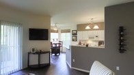 This single-floor residence offers an open-concept layout that effortlessly connects the living, dining, and kitchen areas into one cohesive living space. The living room features rich hardwood-style flooring, a wall-mounted flat-screen television, and neutral-toned walls that create a warm, inviting atmosphere. A ceiling fan overhead adds both function and a casual charm to the space.

The kitchen is well-appointed with white cabinetry, light countertops, and white appliances, including a built-in microwave. A peninsula counter with a raised bar seamlessly separates the kitchen from the living area while maintaining the open feel. An elegant pendant light fixture hangs above, adding a refined touch to the space.

The dining area is bathed in natural light streaming through large windows dressed with warm-toned window treatments, creating a bright and cheerful setting for everyday meals. The dining space flows naturally from both the kitchen and living areas, reinforcing the home's open and connected design.

Throughout the space, the combination of neutral wall tones, consistent flooring, and thoughtful lighting creates a cohesive and comfortable ambiance — ideal for both relaxed everyday living and entertaining guests.