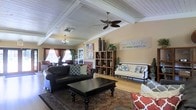 This single-floor space, presented by McKinley, embodies a versatile "Live. Work. Shop. Play." concept, offering a thoughtfully designed interior that balances comfort and functionality. The main living area immediately draws attention with its distinctive beamed ceiling and white-painted wood paneling overhead, lending a classic, cottage-inspired architectural character to the space. A ceiling fan anchors the center of the room, complementing the natural light that filters in through draped windows along the far wall. The warm, neutral-toned walls create an inviting ambiance throughout. The living area is furnished with a rich, dark leather sofa and an elegant upholstered bench, arranged around a bold, dark-stained coffee table set atop a traditional patterned area rug over light hardwood flooring. A wooden open-shelf bookcase serves as a stylish room divider and display unit, adding depth and visual interest to the layout. A chandelier visible in the adjoining area hints at additional well-appointed spaces beyond the main room. Decorative touches, including tall vases, potted greenery, and patterned throw pillows, contribute to the space's warm and curated aesthetic, making it an appealing and multi-purpose environment.